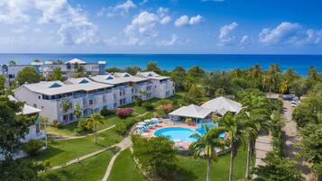 Relax by the resort's central pool surrounded by lush greenery and palm trees, with ocean views from the balconies of multiple buildings.