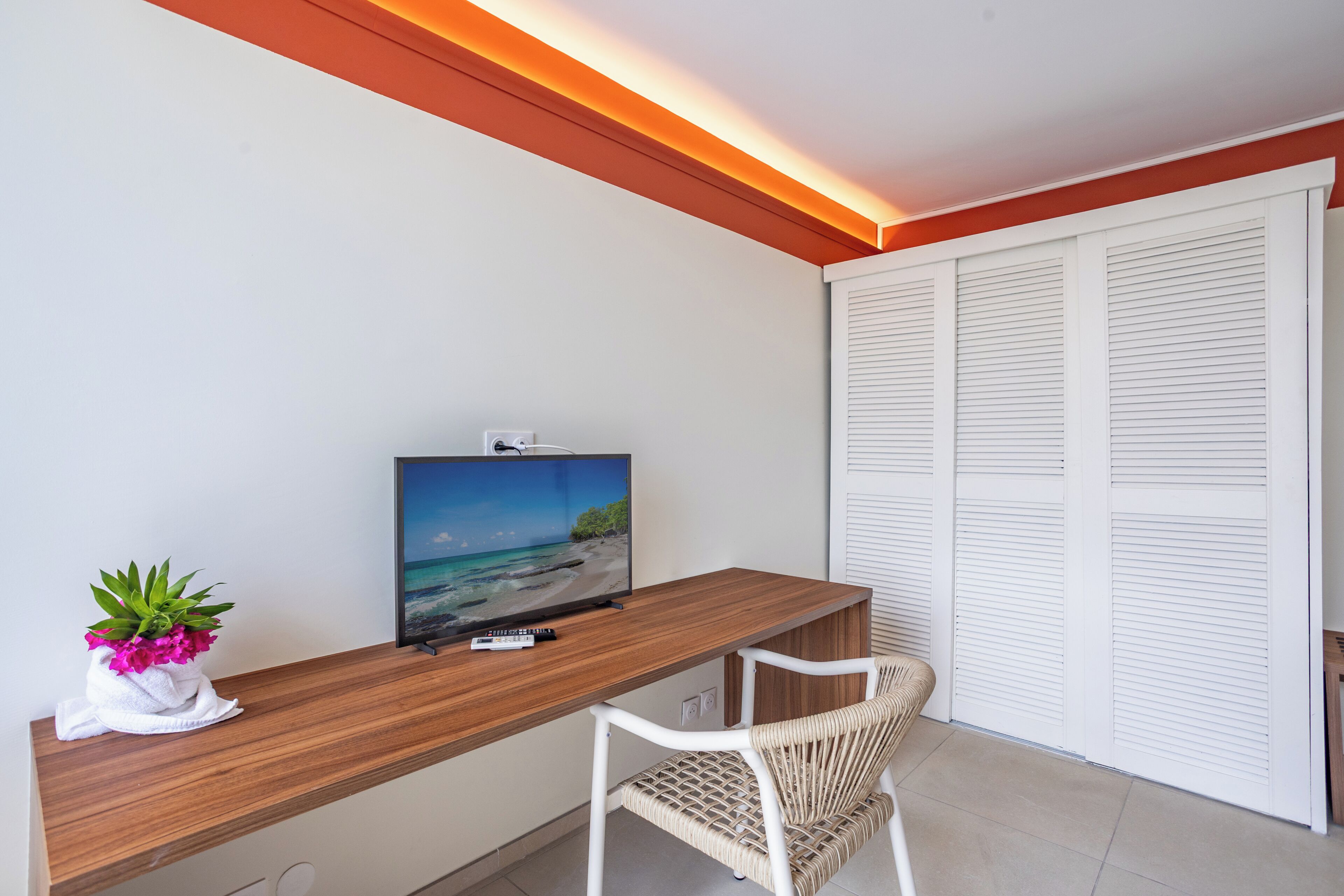 Una habitación con un escritorio de madera, un televisor mostrando una escena de playa, una silla blanca y un armario blanco con puertas a listones.