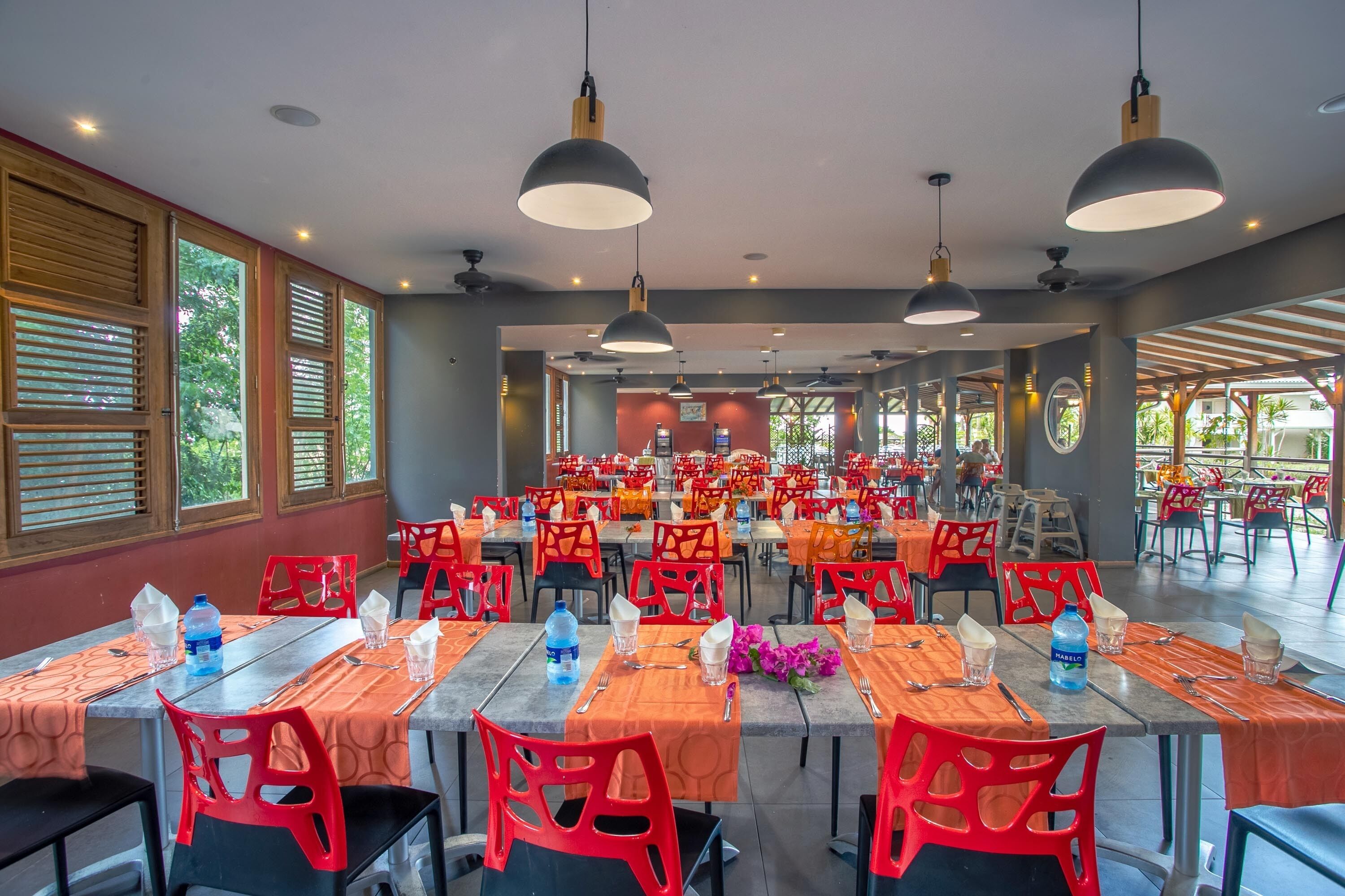 A spacious dining area with multiple tables and chairs, large pendant lights, and windows with wooden shutters.