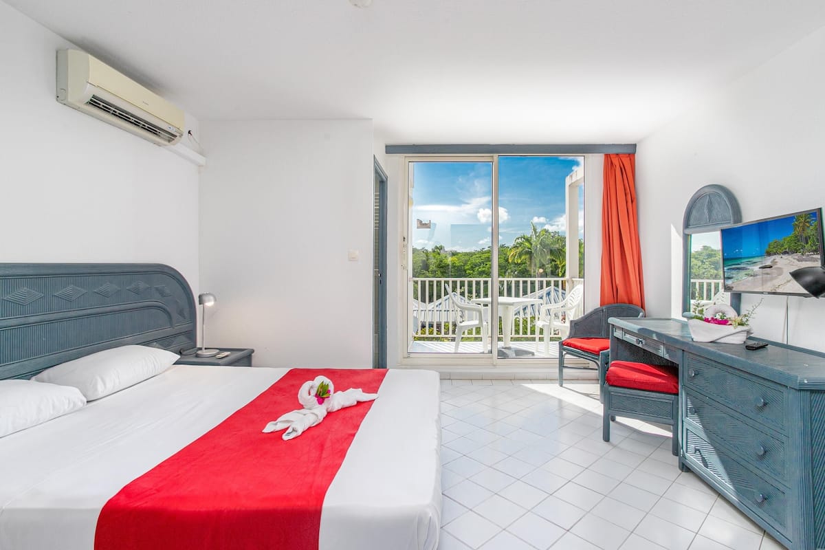 A hotel room with a bed, a desk, a chair, a TV, and a balcony with a view of greenery.