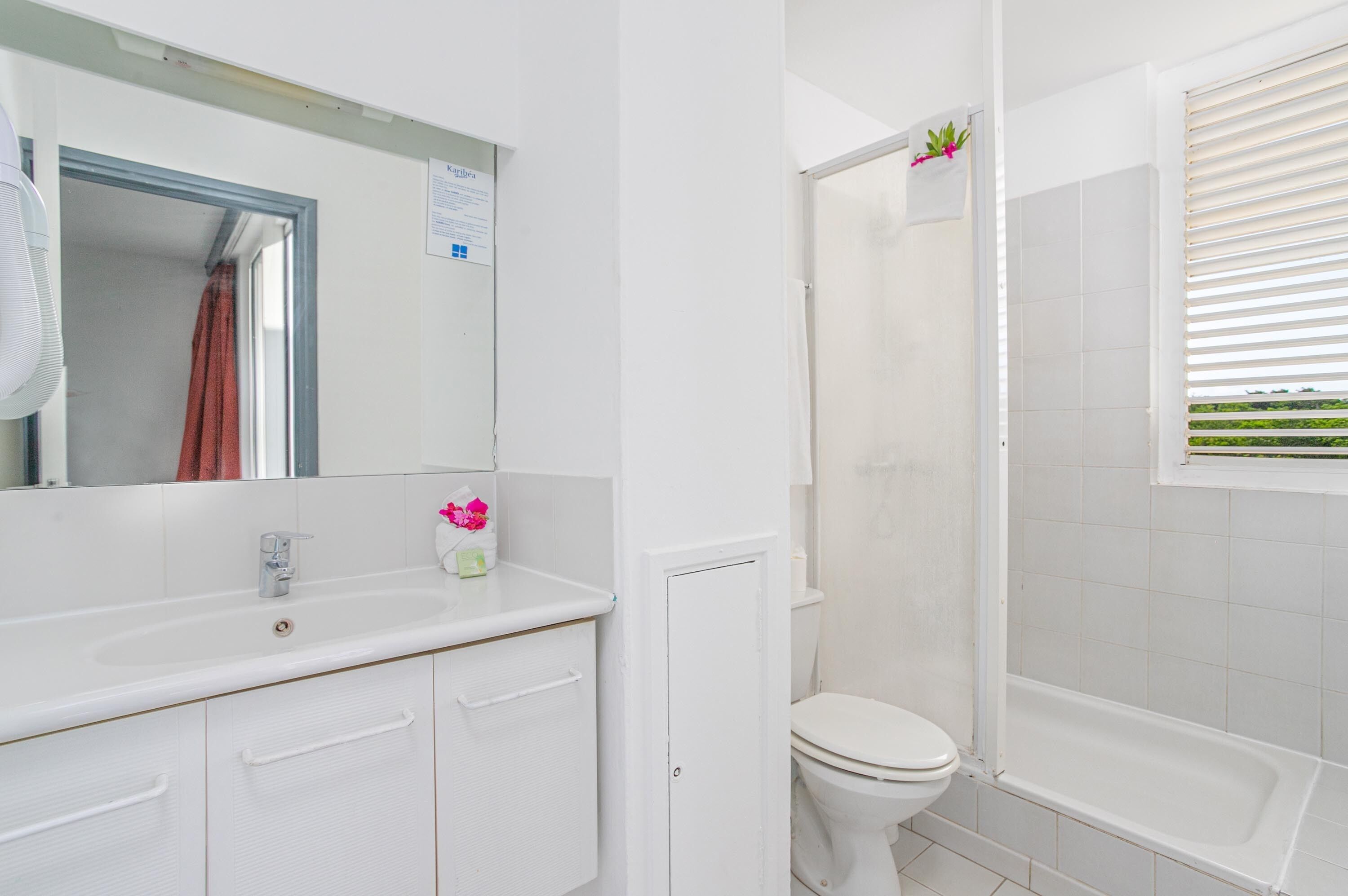Un baño con lavamanos blanco, espejo y retrete. Hay una ventana con persianas y una toalla colgada en la pared.