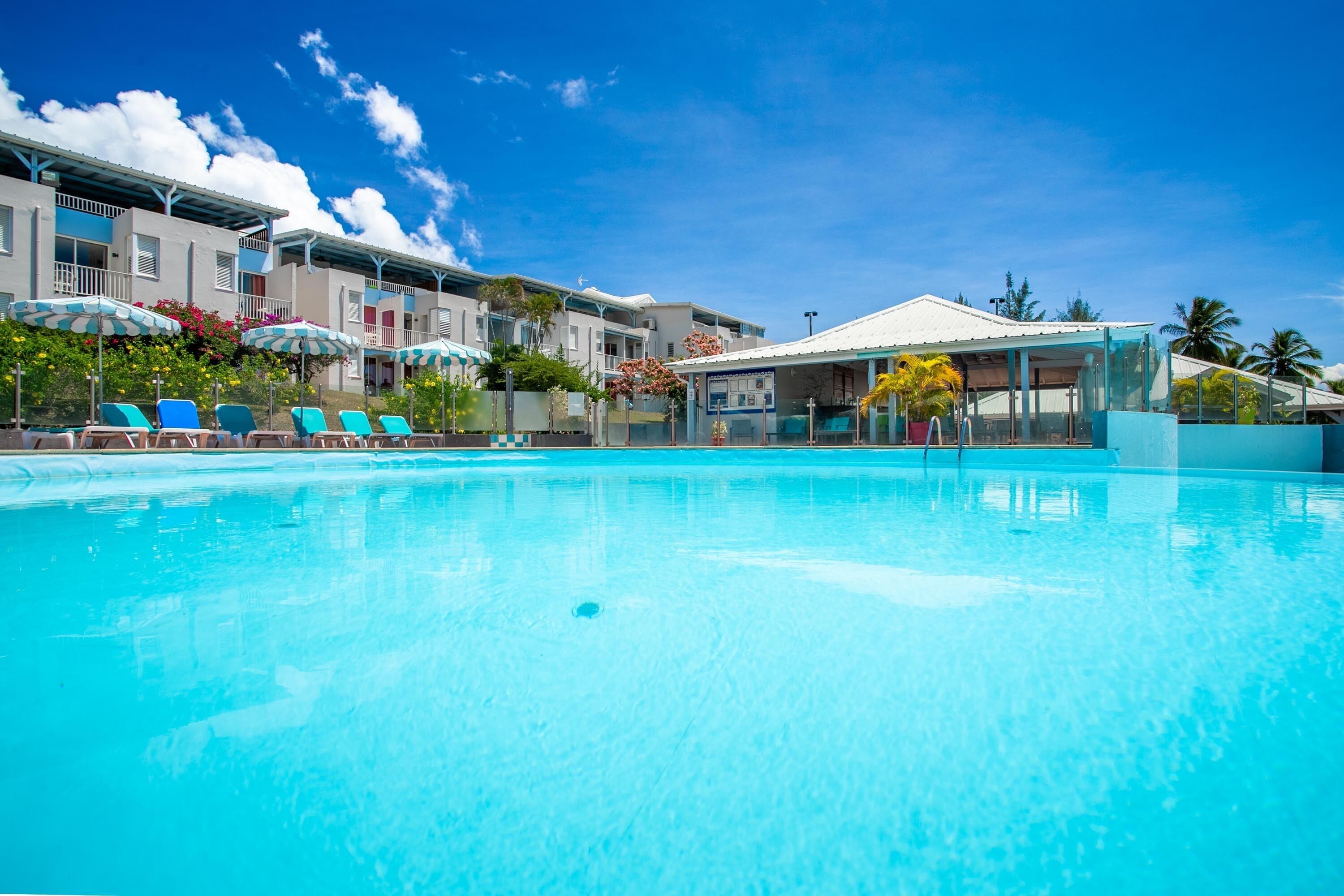 A swimming pool with lounge chairs and umbrellas, surrounded by multi-story buildings with balconies and a covered area.