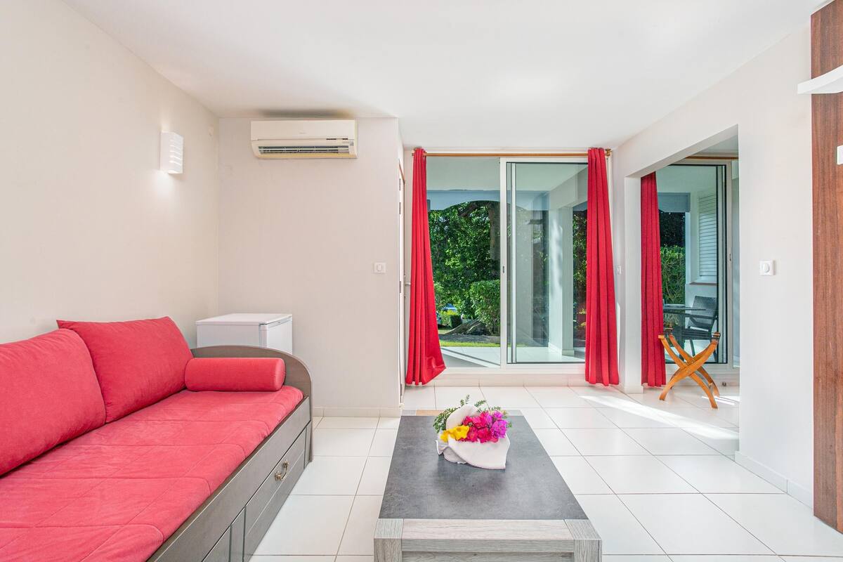 A modern living room with a red sofa, a small table with a flower arrangement, and a view of a garden through a sliding glass door.
