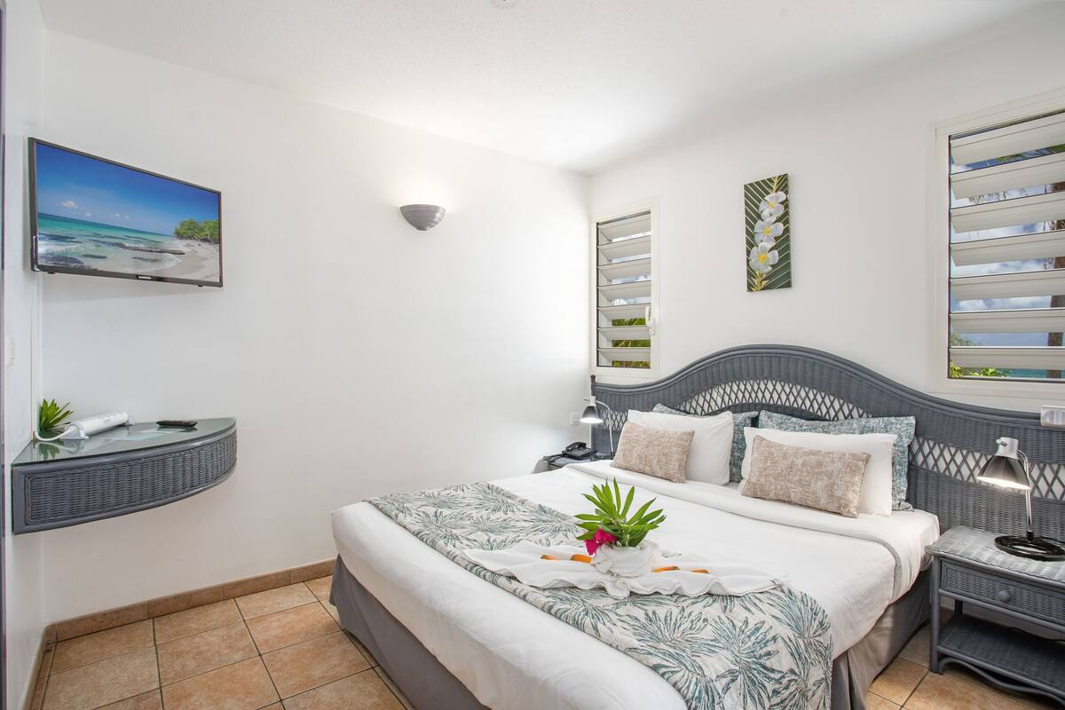 A bedroom with a bed, a TV, a wall-mounted shelf, and a window with blinds.