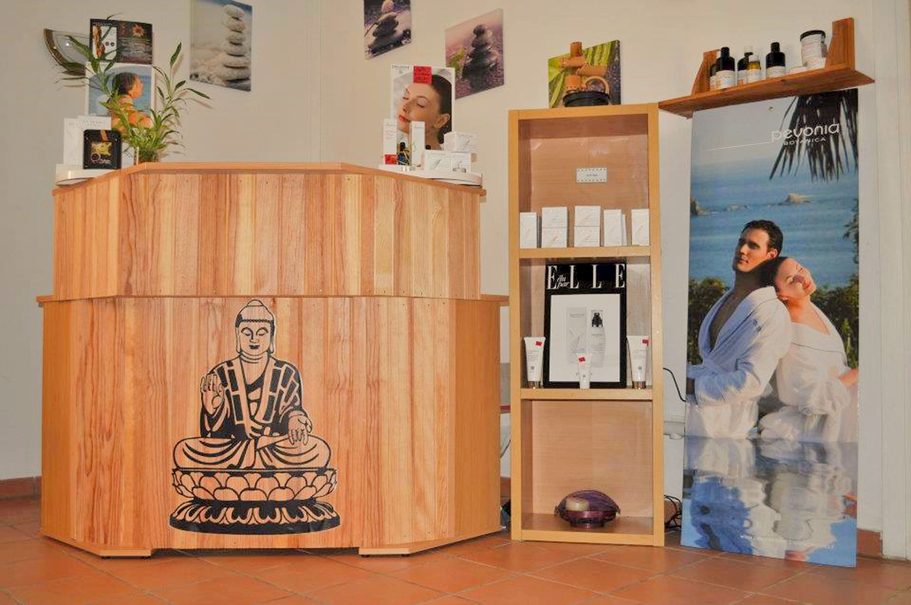 Un comptoir d’accueil en bois orné d’une statuette évoquant la méditation, une étagère avec des produits de soins pour la peau et une affiche représentant un couple au bord de la mer.