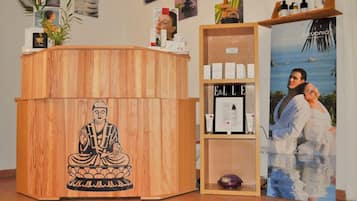 Un comptoir d’accueil en bois orné d’une statuette évoquant la méditation, une étagère avec des produits de soins pour la peau et une affiche représentant un couple au bord de la mer.