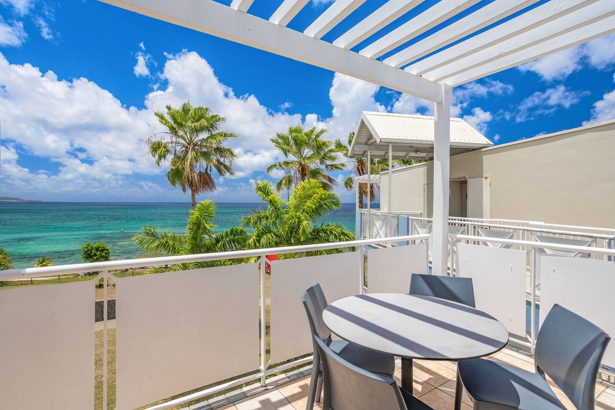 A balcony with a table and chairs, overlooking a beach with palm trees and the ocean.