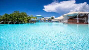 A clear blue pool with lounge chairs and umbrellas, surrounded by lush greenery and a covered pavilion.