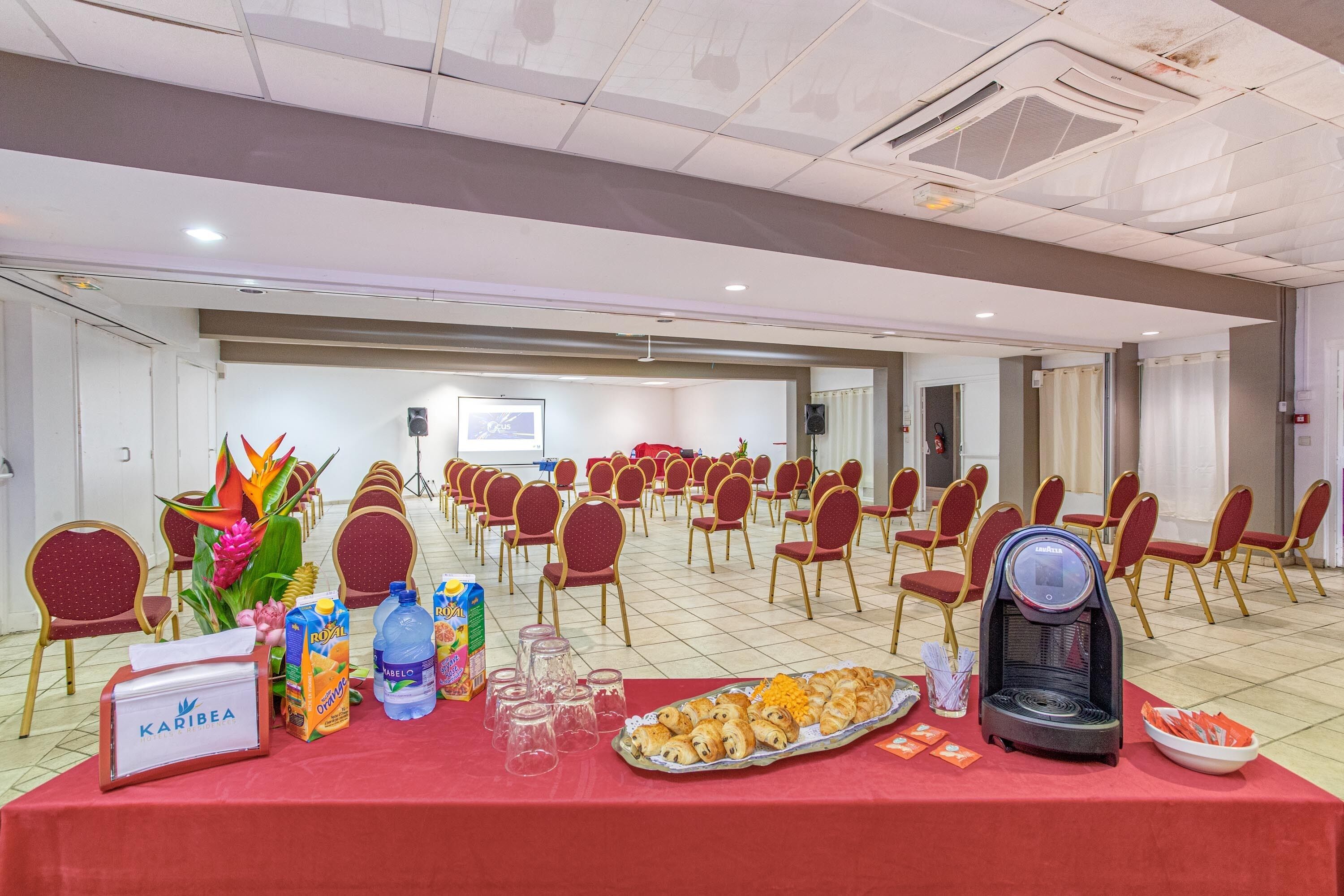 A conference room with rows of red chairs, a table with refreshments, and a projector screen in the background.