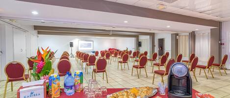 A conference room with rows of red chairs, a table with refreshments, and a projector screen in the background.
