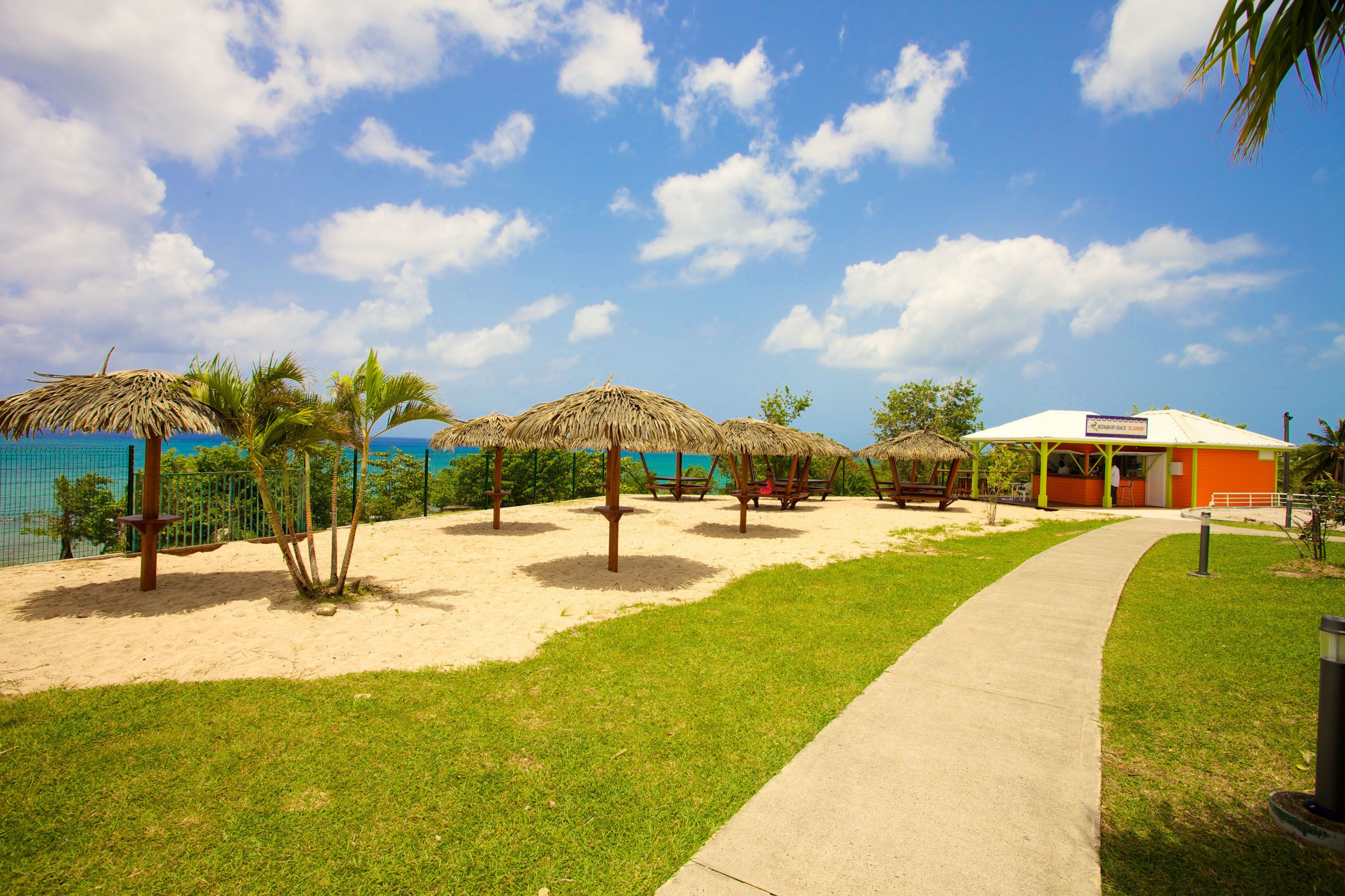 A beachside area with thatched umbrellas, a concrete walkway, and a building with a sign.