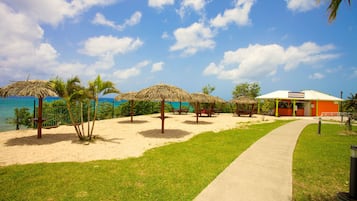 A beachside area with thatched umbrellas, a concrete walkway, and a building with a sign.