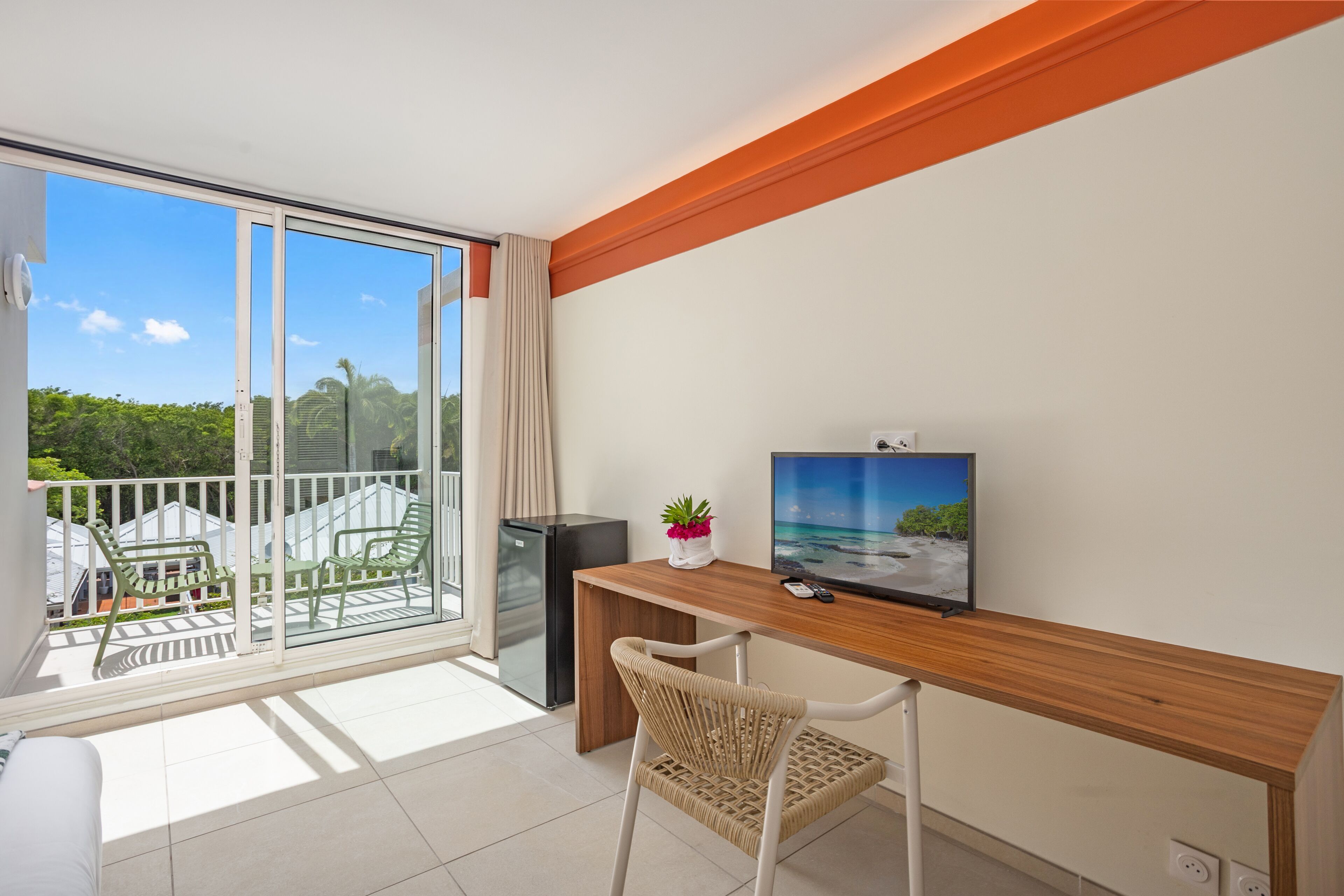 A balcony with a view of greenery and a building, a wooden desk with a television, and a wicker chair.