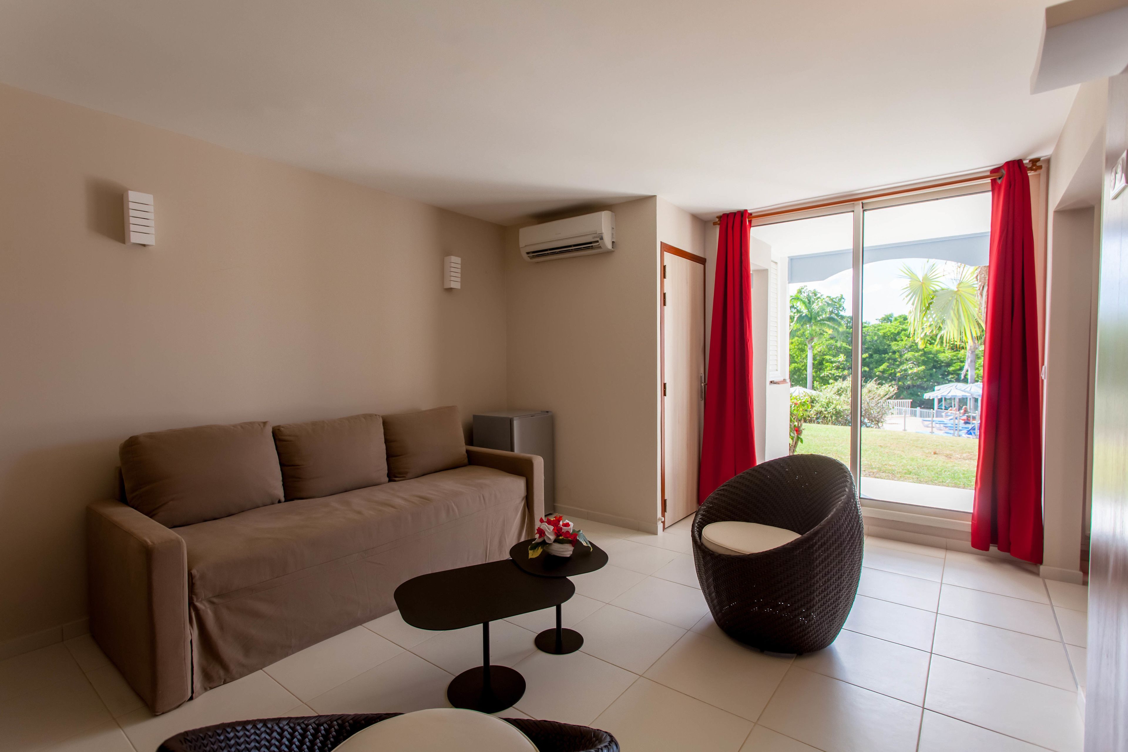 A modern living room with a sofa, a round wicker chair, a small table, and a sliding glass door leading to a balcony with a view of greenery.