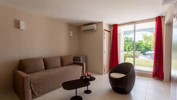 A modern living room with a sofa, a round wicker chair, a small table, and a sliding glass door leading to a balcony with a view of greenery.