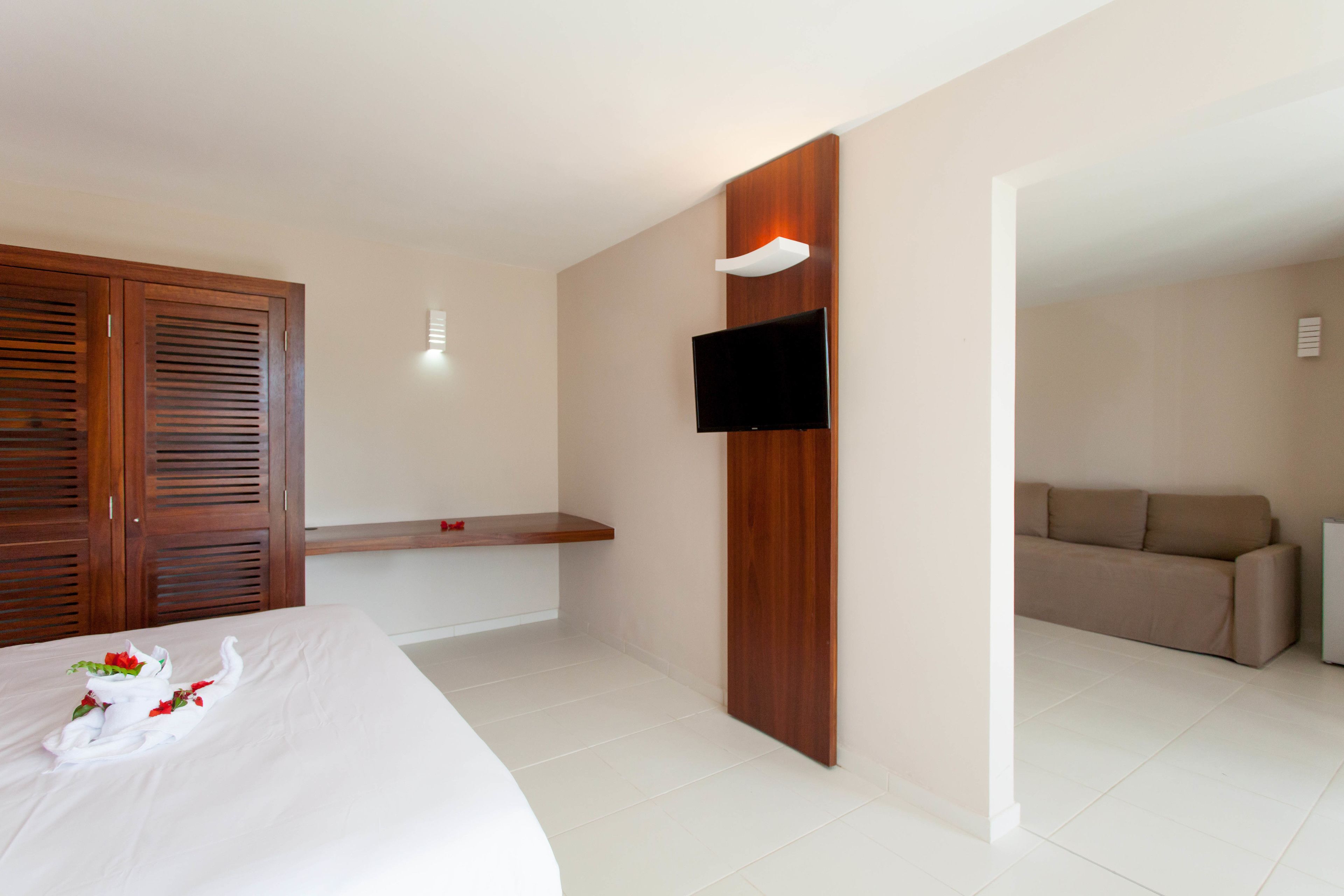 A hotel room with a bed, a wooden wardrobe, a mounted TV, and a sofa in the adjacent living area.