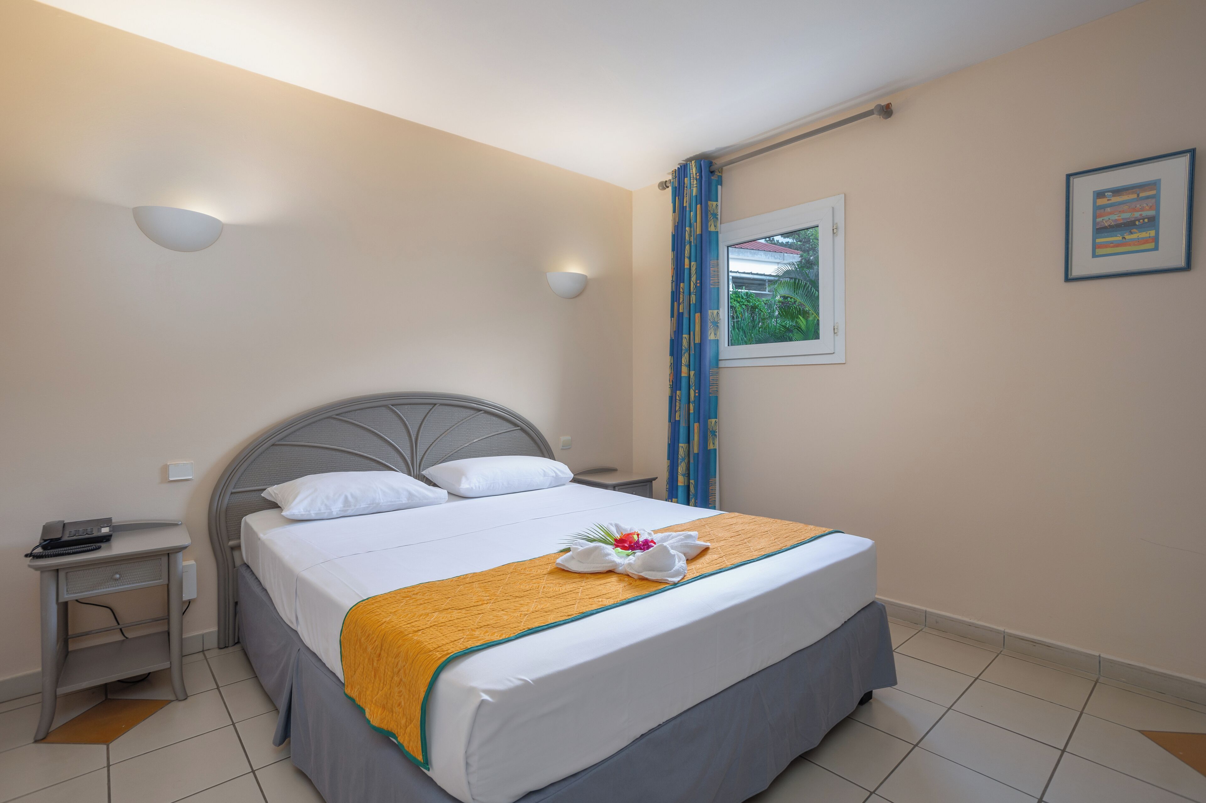A neatly made bed with a white comforter and a yellow accent, a bedside table with a phone, and a window with blue curtains.