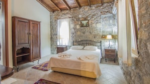 A bedroom with a stone wall, wooden ceiling, a bed with white linens, a wooden wardrobe, a nightstand with a lamp, and a window with curtains. - Doga Apartments (Fethiye)