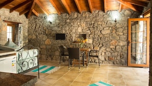 A rustic stone-walled room with a wooden ceiling, a dining table with chairs, a stone fireplace, and a large window. - Doga Apartments (Fethiye)
