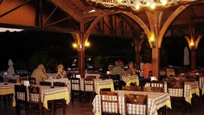 An outdoor dining area with tables covered in checkered tablecloths, wooden chairs, and a rustic wooden ceiling structure. - Candan Beach Hotel (Marmaris)