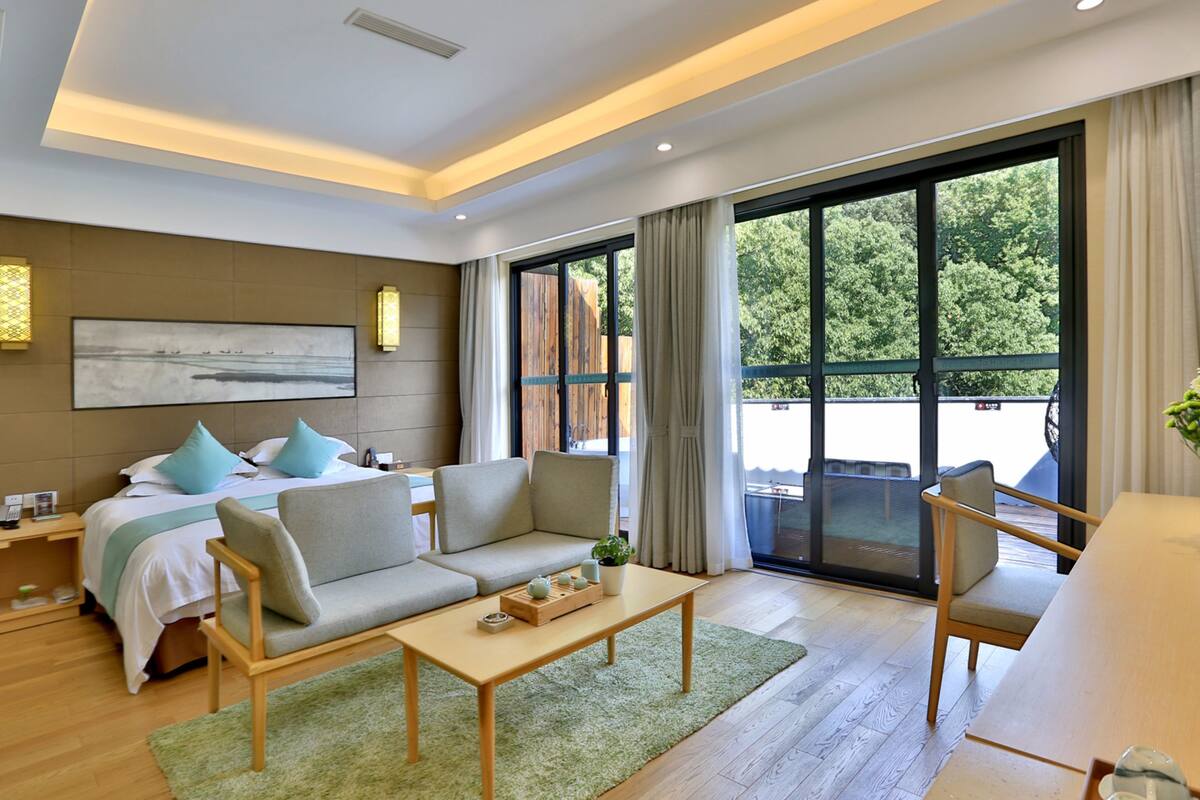 A modern hotel room with a bed, sofa, armchair, and a dining table. There is a large window with a view of greenery outside.