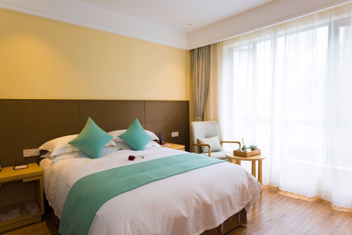 A hotel room with a large bed, two pillows, a bedside table, a chair, and a window with curtains.