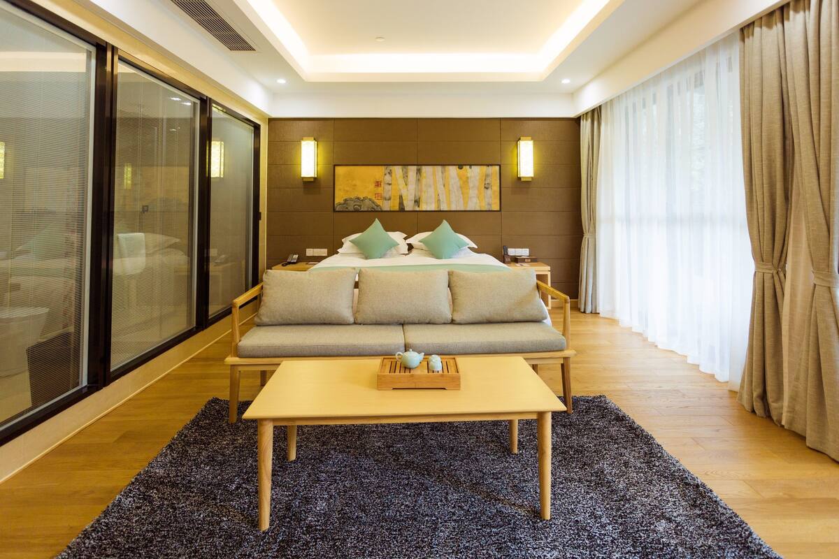 A modern hotel room with a large bed, a sofa, a coffee table, and a sliding glass door.