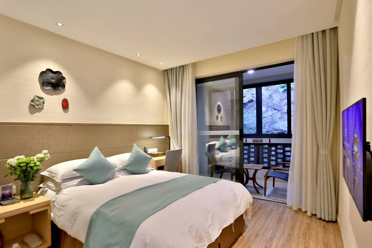 A hotel room with a bed, a nightstand, a vase with flowers, a desk with a chair, and a view of the outdoors through a sliding door.