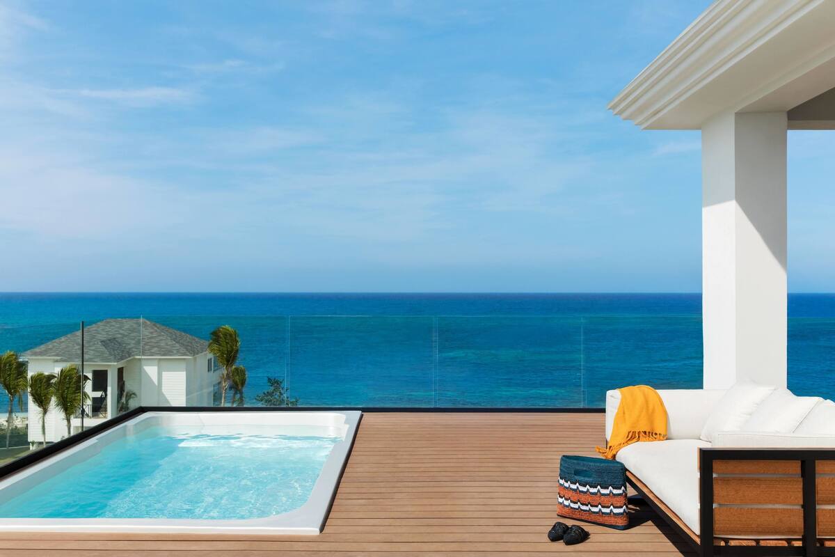 Eine Holzterrasse mit Whirlpool und einem weißen Sofa, ausgestattet mit einem gelben Überwurf, mit Blick auf das klare, blaue Meer.