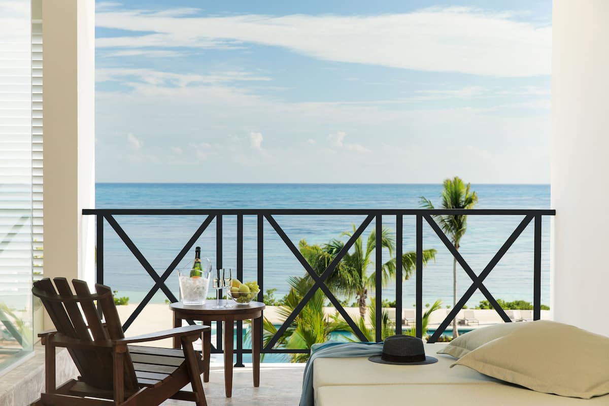Ein Balkon mit Blick auf den Ozean, ein Tisch mit Getränken, ein Stuhl und ein Bett mit Kissen.