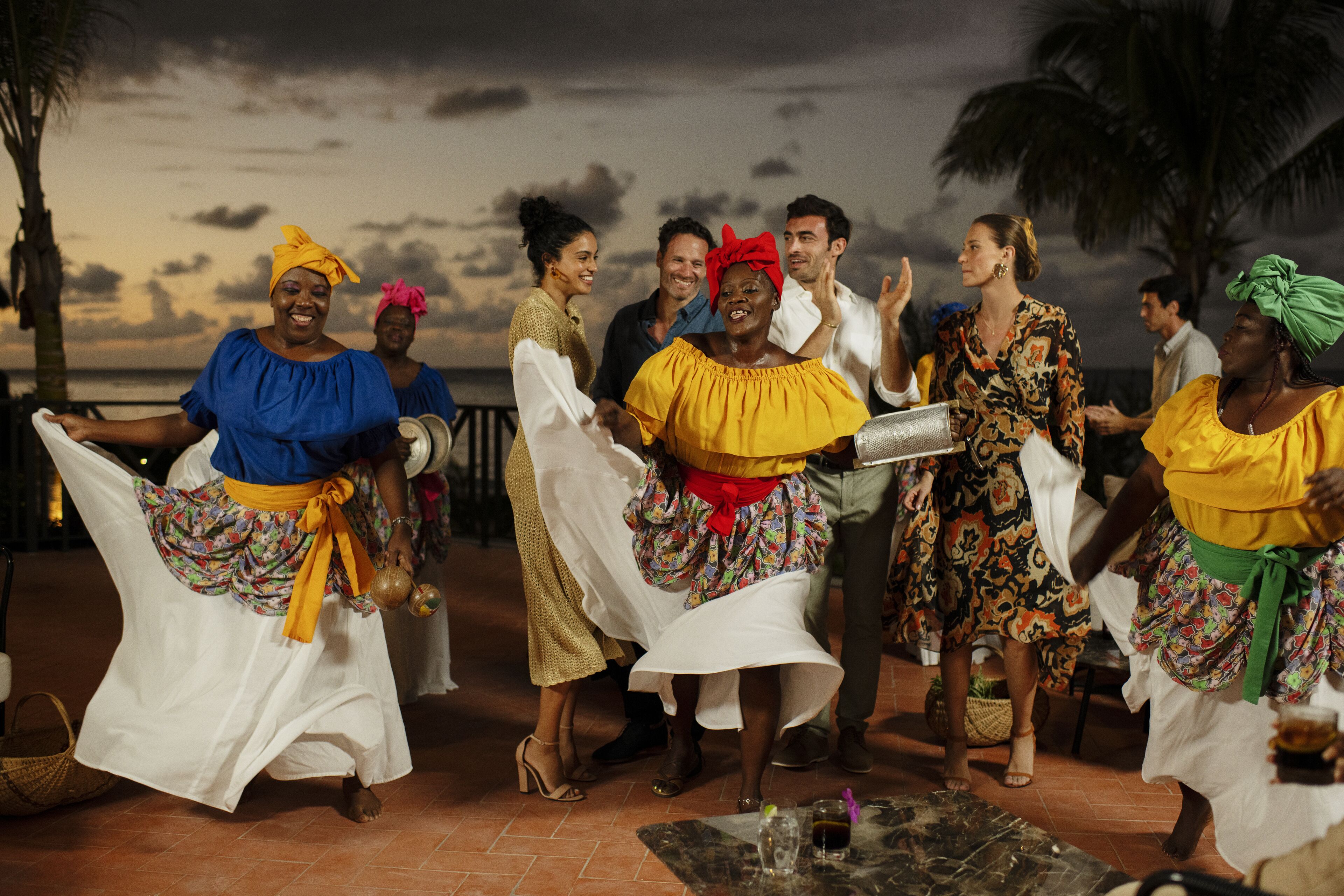 A group of people dancing on a terrace with a view of the ocean and palm trees.
