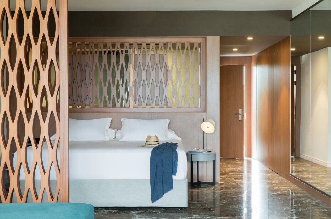 A modern hotel room with a bed, a nightstand, a lamp, and a decorative wooden partition.