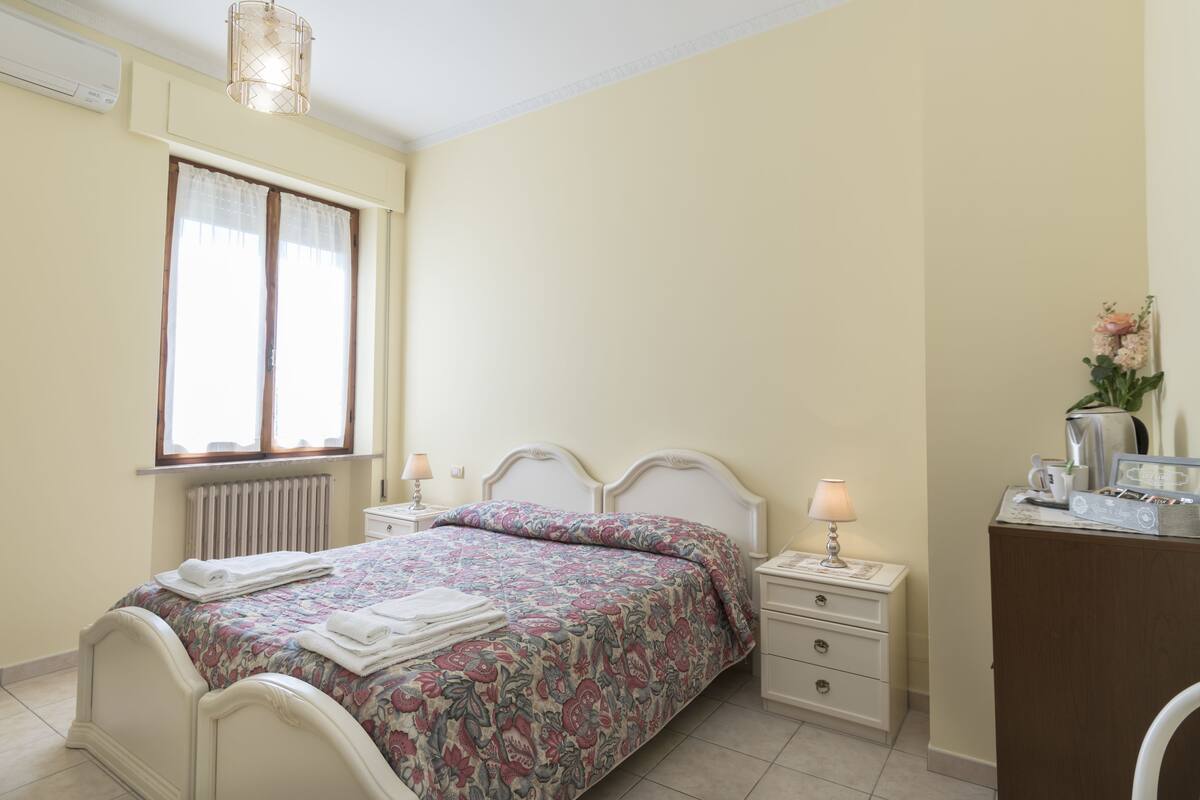 A bedroom with a bed, bedside tables, a dresser, a radiator, and a window with curtains.