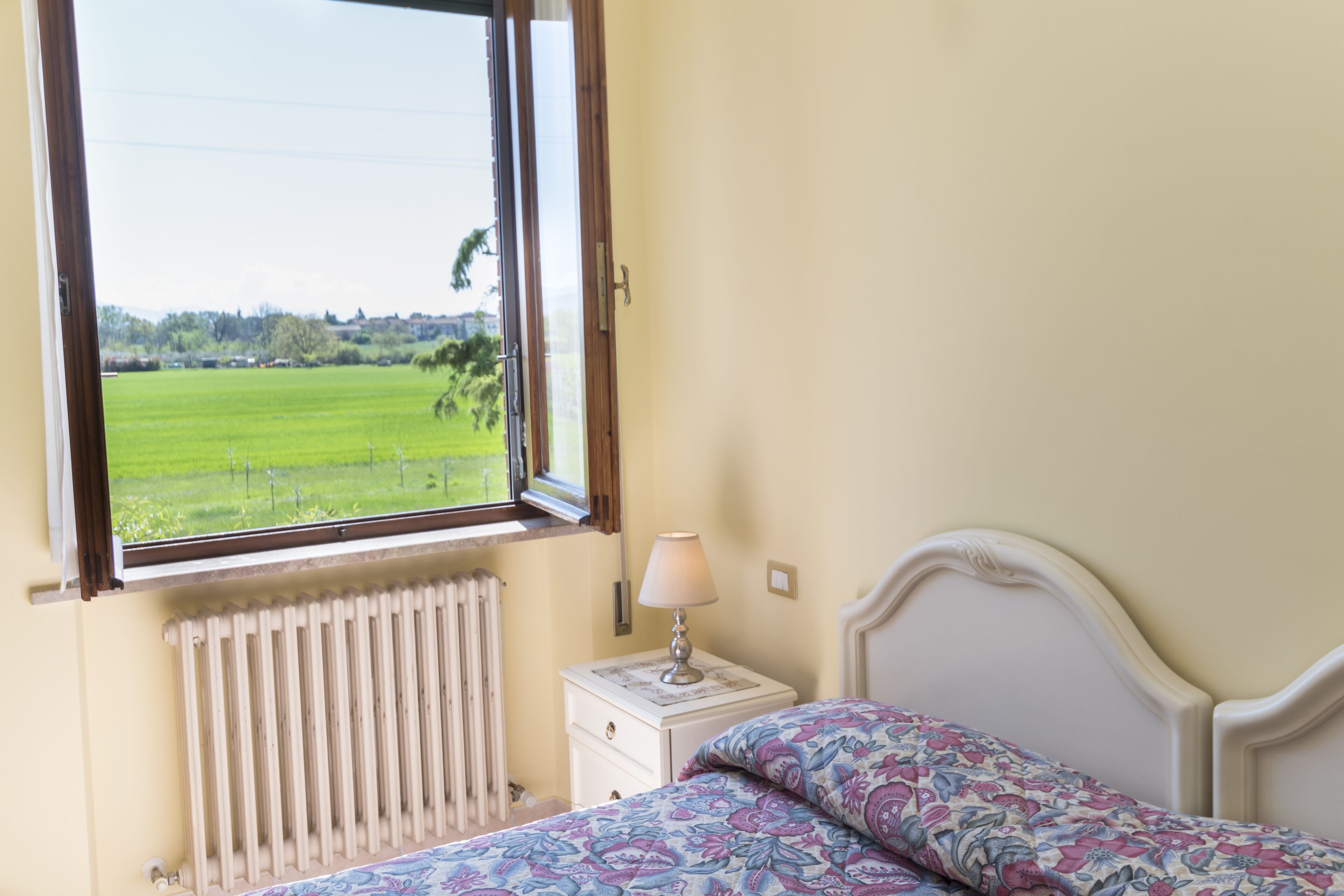 Une chambre à coucher comprenant un lit, une table de chevet, une lampe, une fenêtre donnant sur un champ verdoyant et un radiateur.
