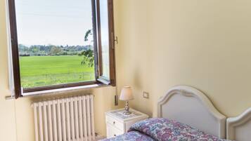 Une chambre à coucher comprenant un lit, une table de chevet, une lampe, une fenêtre donnant sur un champ verdoyant et un radiateur.