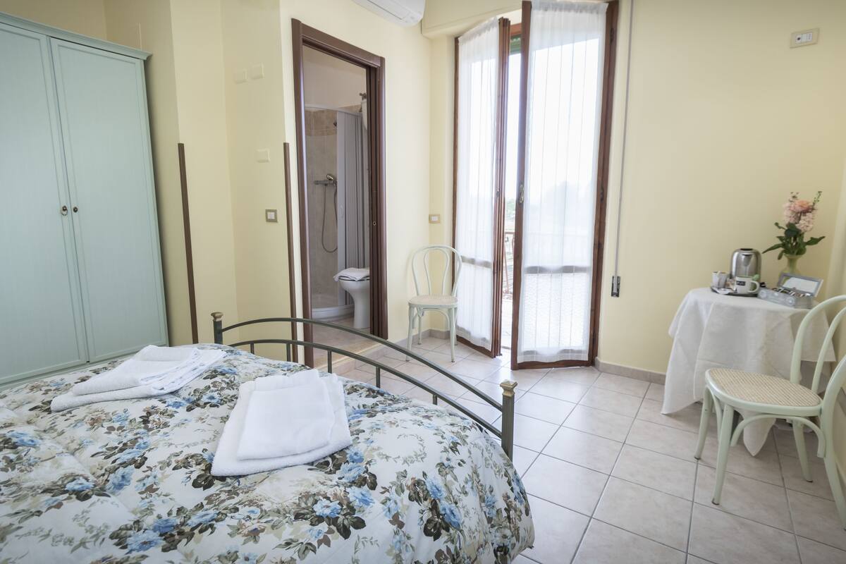 A bedroom with a bed, a wardrobe, a bathroom with a shower, and a table with a chair.