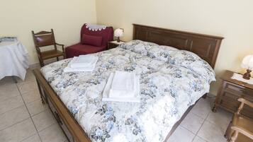 Una cama bien hecha con ropa de cama floral, una mesita de noche de madera con lámpara, una silla roja y una pequeña mesa con mantel blanco.