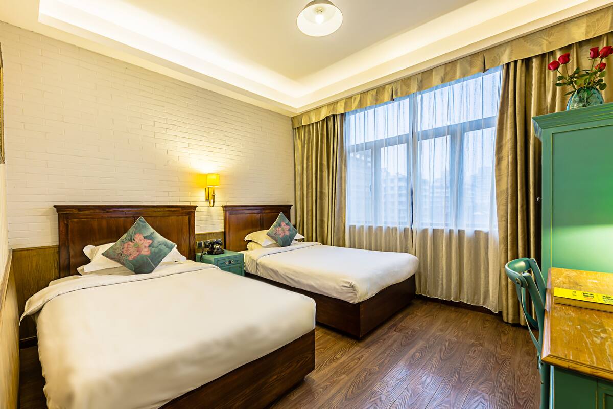 A hotel room with two beds, a wooden headboard, a nightstand, a lamp, a desk, and a window with curtains.
