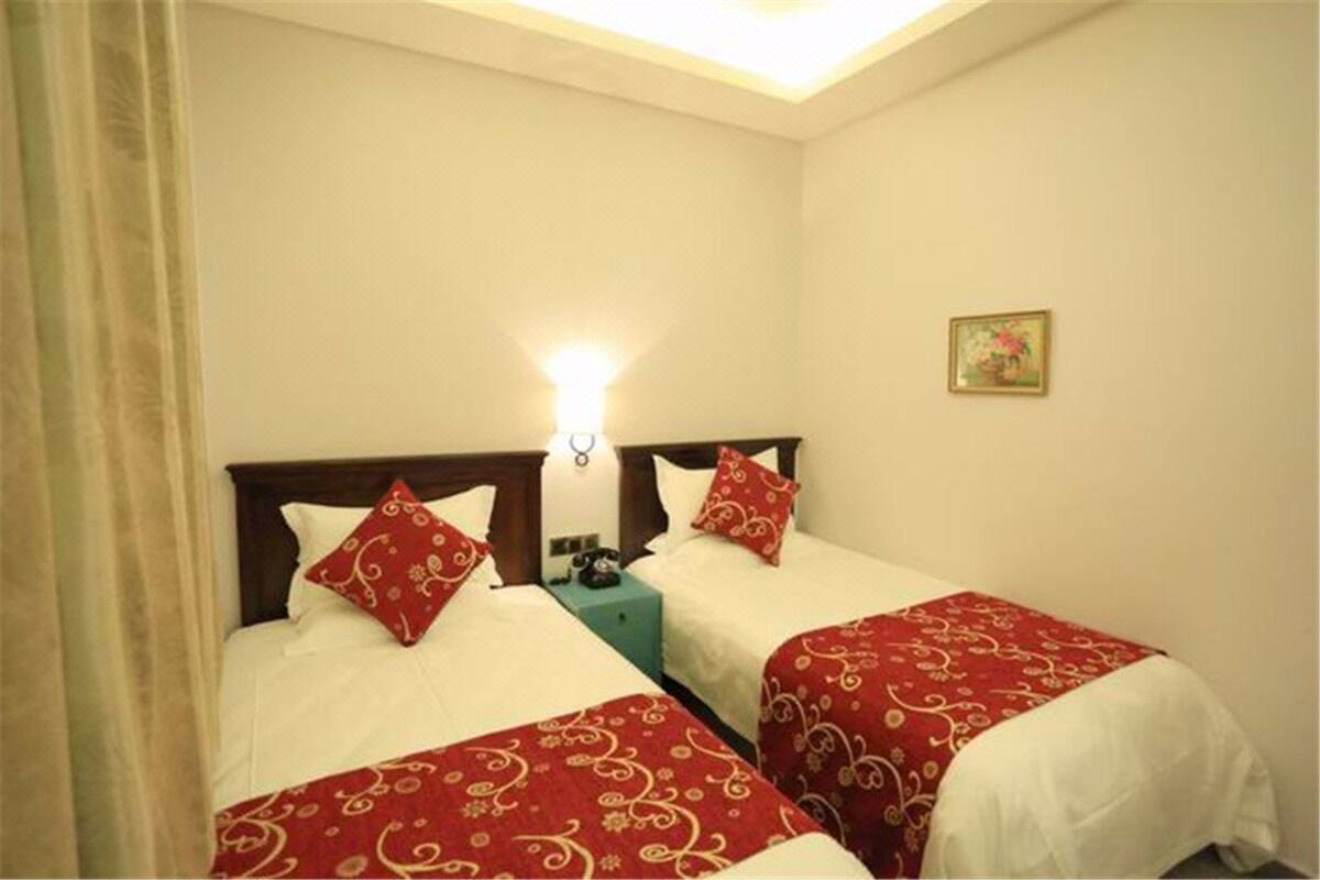 A hotel room with two beds, each with a red and white bedspread, a bedside table with a lamp, and a small painting on the wall.