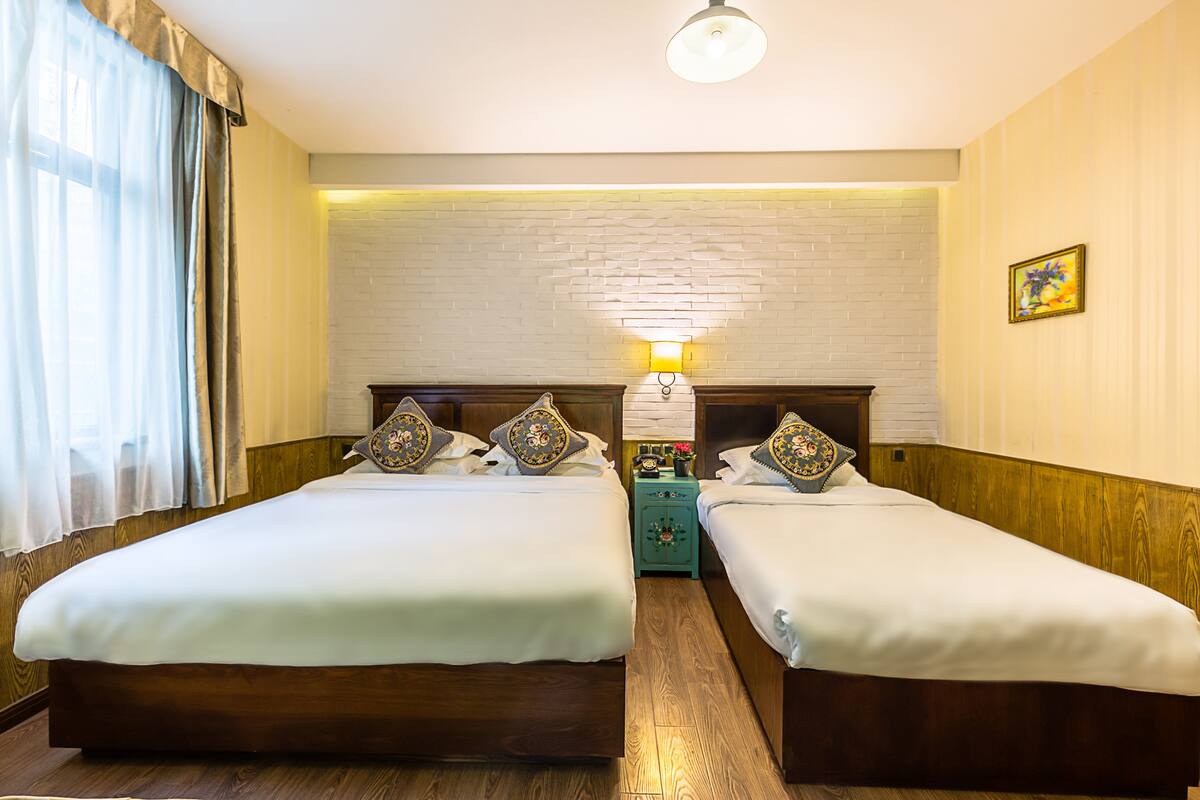 A hotel room with two beds, a brick wall, a nightstand with a lamp, and a painting on the wall.