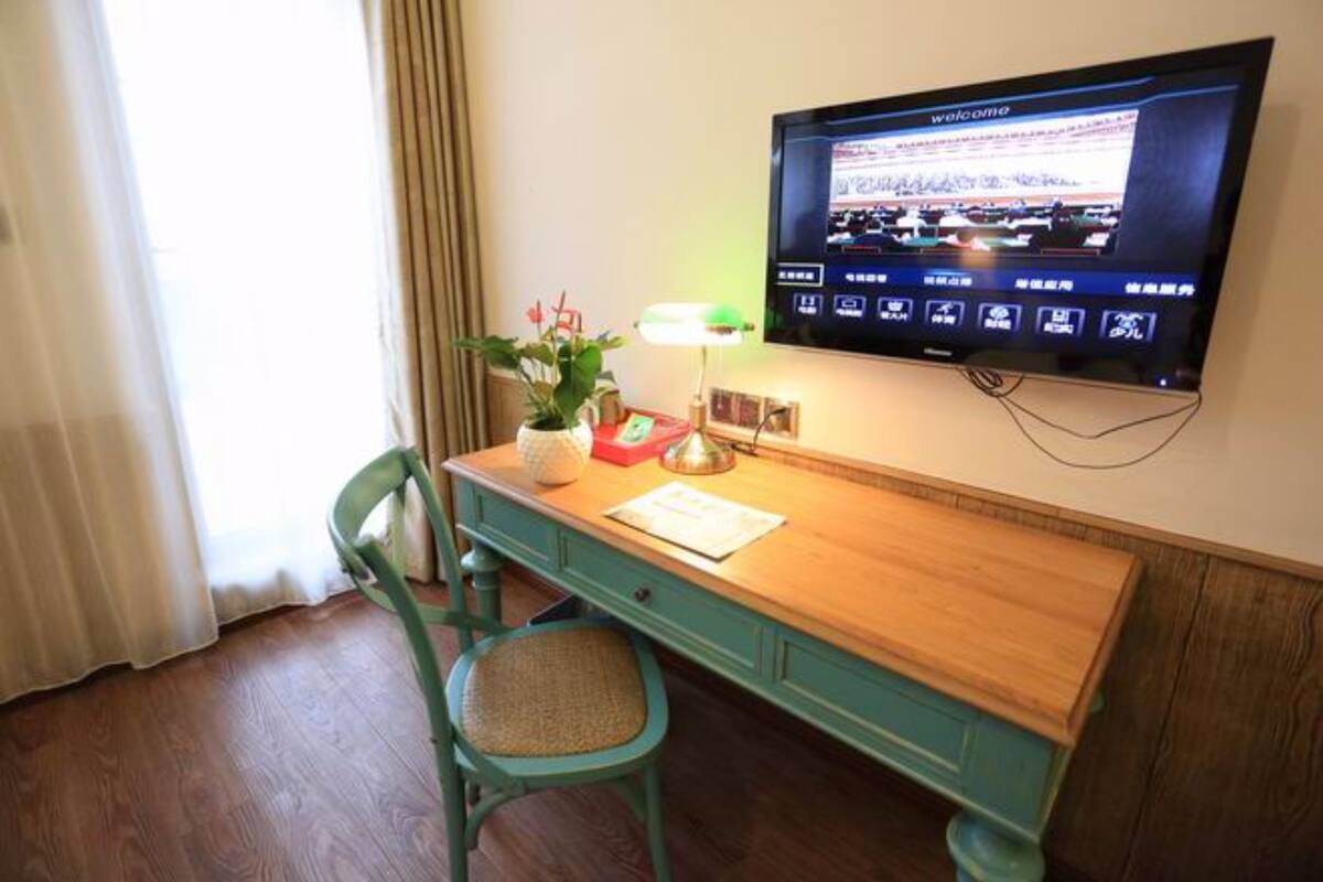 A hotel room with a wooden desk, a green chair, a flat-screen TV mounted on the wall, and a window with curtains.