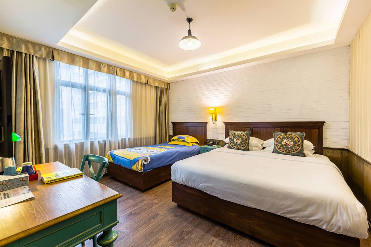 A hotel room with two beds, a desk, a chair, a lamp, and a window with curtains.