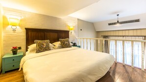 A bedroom with a large bed, two bedside tables, a nightstand, a brick wall, and a balcony with a railing.