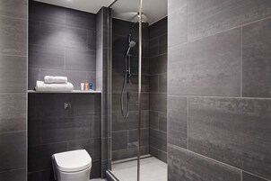 Un baño moderno con azulejos oscuros, ducha con mampara de vidrio, inodoro y repisa con toallas y artículos de aseo.