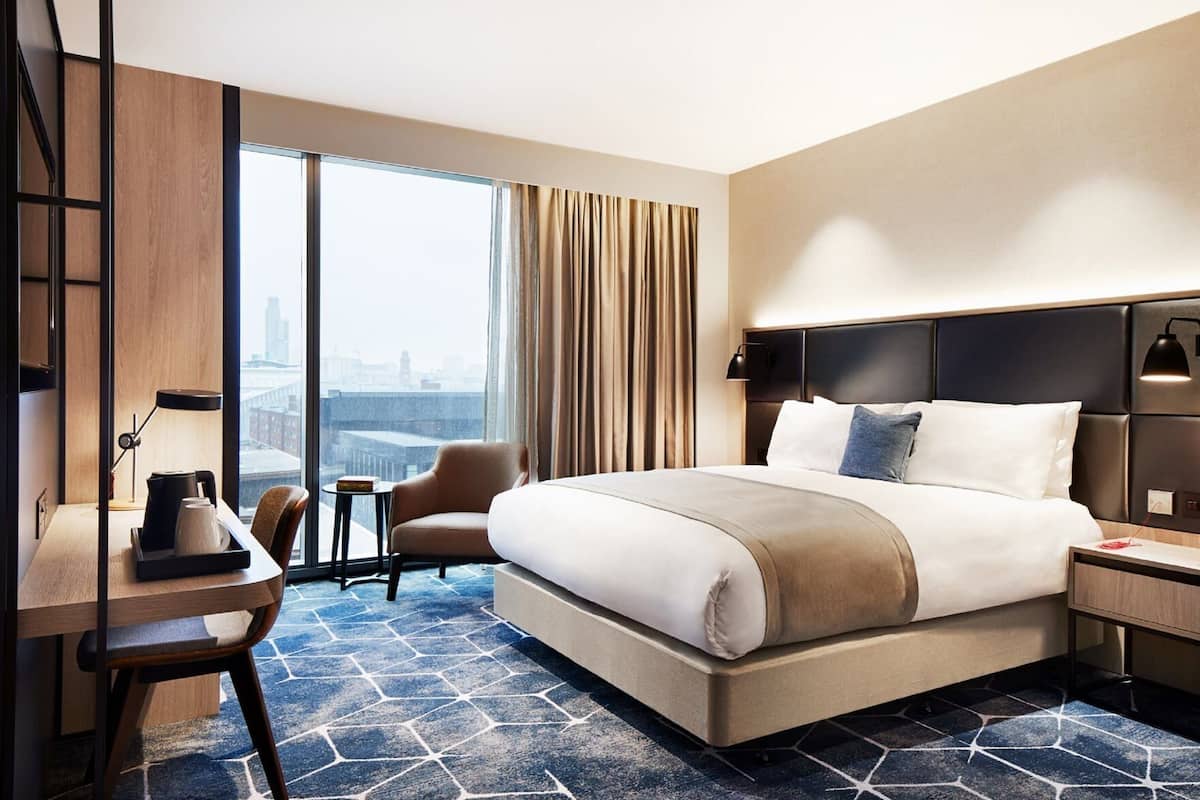 Ein Hotelzimmer mit einem großen Bett, einem Schreibtisch, einem Stuhl, einem kleinen Tisch und Blick auf die Stadt.