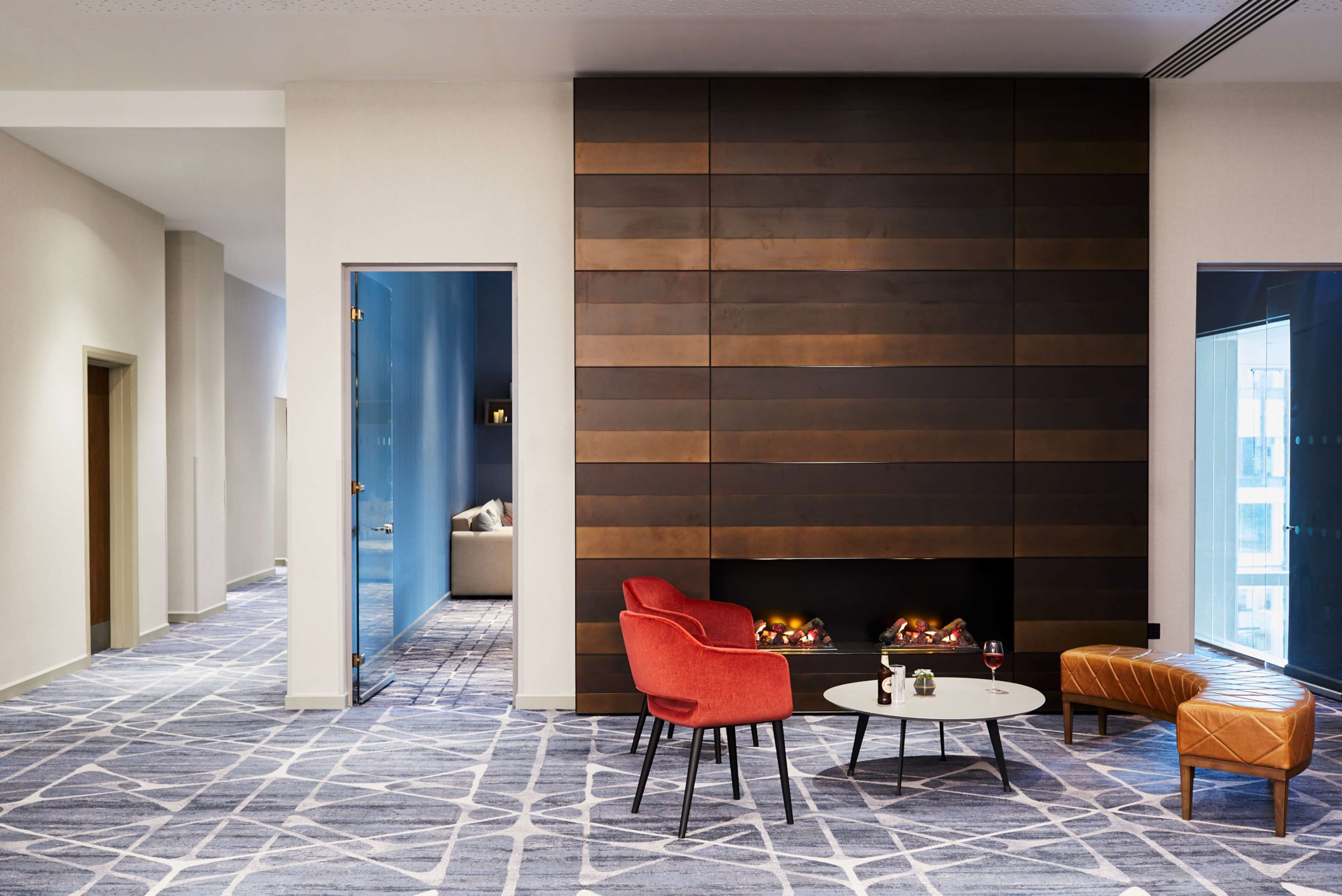 Un hall d’hôtel moderne avec un tapis à motifs, une cheminée, des fauteuils rouges et orange, et une vue sur l’extérieur à travers une porte vitrée.