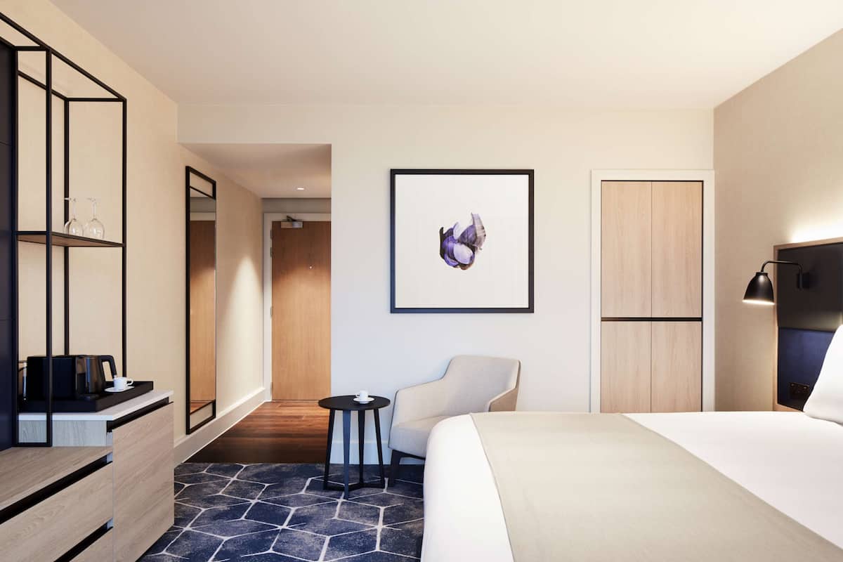 Ein modernes Hotelzimmer mit einem Bett, einem Sessel, einem kleinen Tisch, einer Kaffeemaschine, einem Spiegel und einem gerahmten Kunstwerk an der Wand.