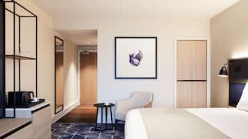A modern hotel room with a bed, a chair, a small table, a coffee machine, a mirror, and a framed artwork on the wall.