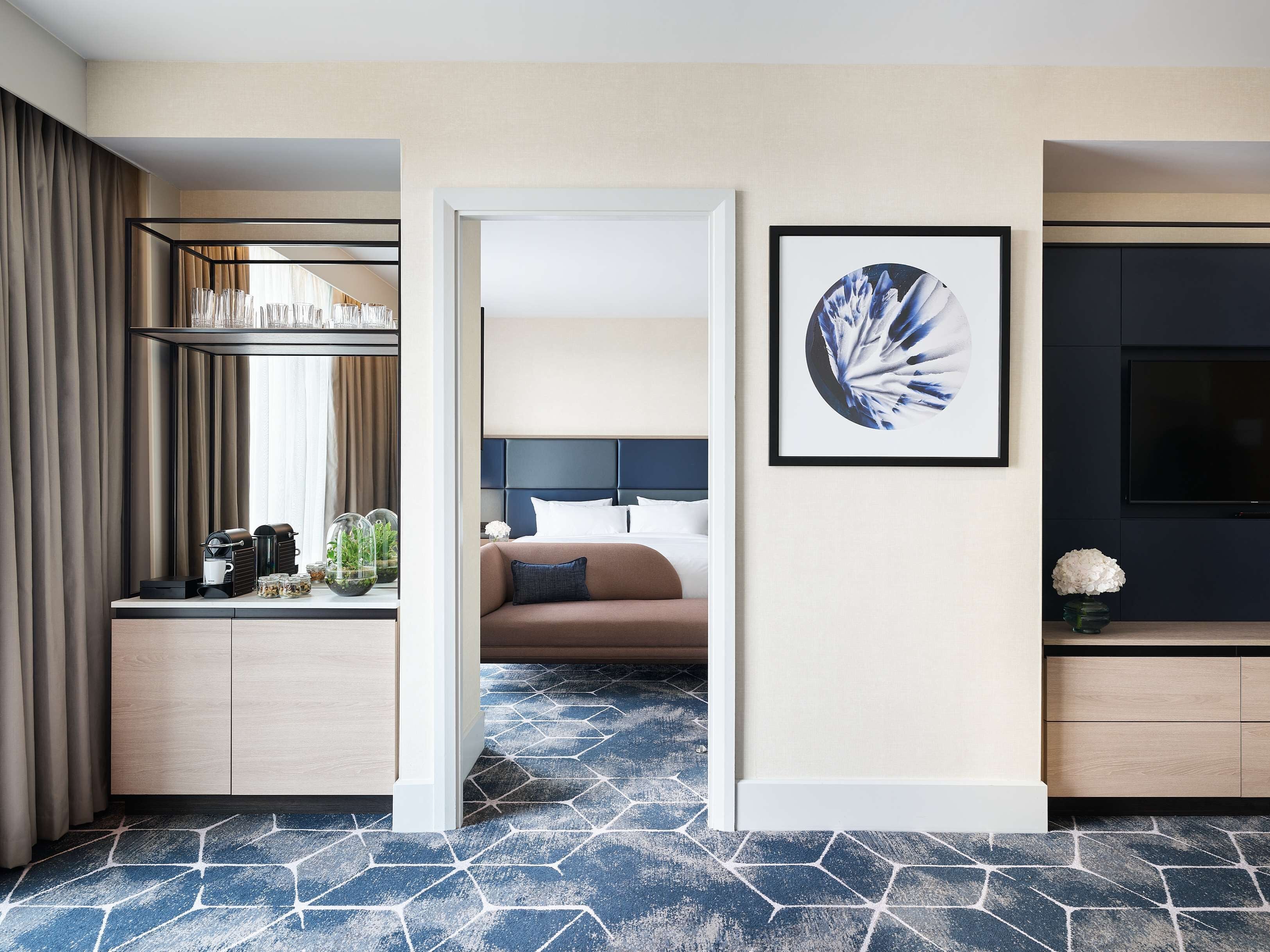 A hotel room with a bed, a desk with a coffee maker, a TV, and a framed artwork on the wall.