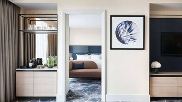A hotel room with a bed, a desk with a coffee maker, a TV, and a framed artwork on the wall.